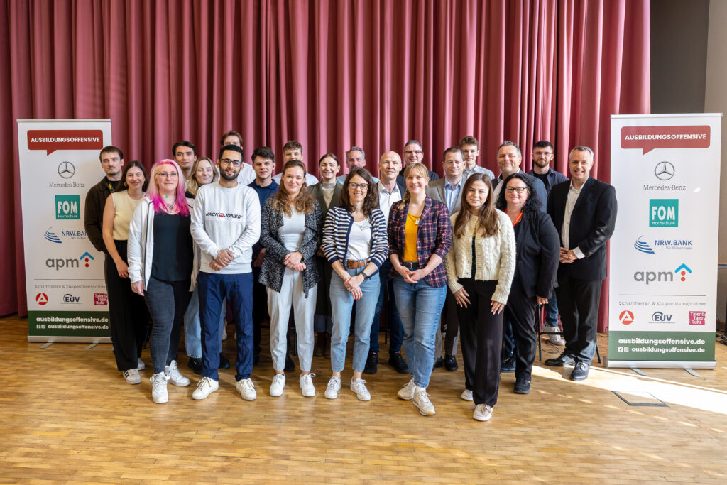 Gruppenfoto von Teilnehmenden und OrganisatorInnen der Ausbildungsoffensive vor einem roten Vorhang. Die bunt gemischte Gruppe aus jungen Auszubildenden, UnternehmensvertreterInnen und Projektbeteiligten steht zwischen zwei Roll-ups mit den Logos der Partner wie Mercedes-Benz, FOM Hochschule, NRW.BANK und apm.
