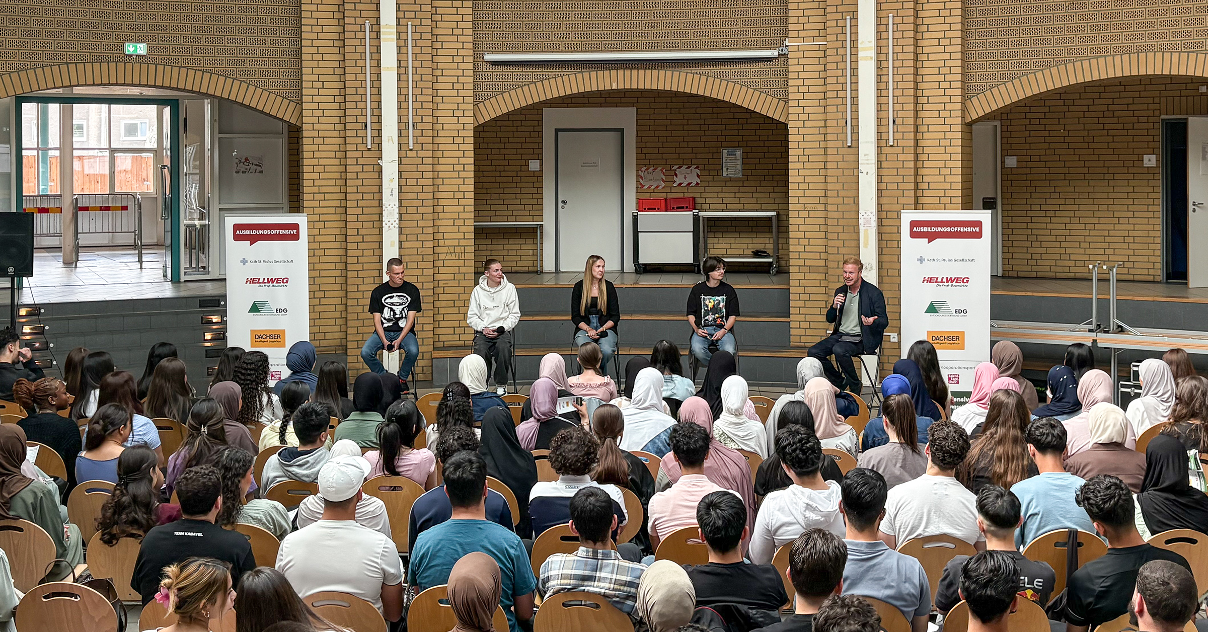 Sechs junge Menschen sitzen bei einer Talkrunde der Ausbildungsoffensive auf einer Bühne in einer Schulaula. Sie berichten vor einem großen, vielfältigen Schülerpublikum über ihre Ausbildungserfahrungen. Zwei Roll-ups mit Logos von Ausbildungsunternehmen wie Hellweg, EDG und DACHSER flankieren die Bühne.