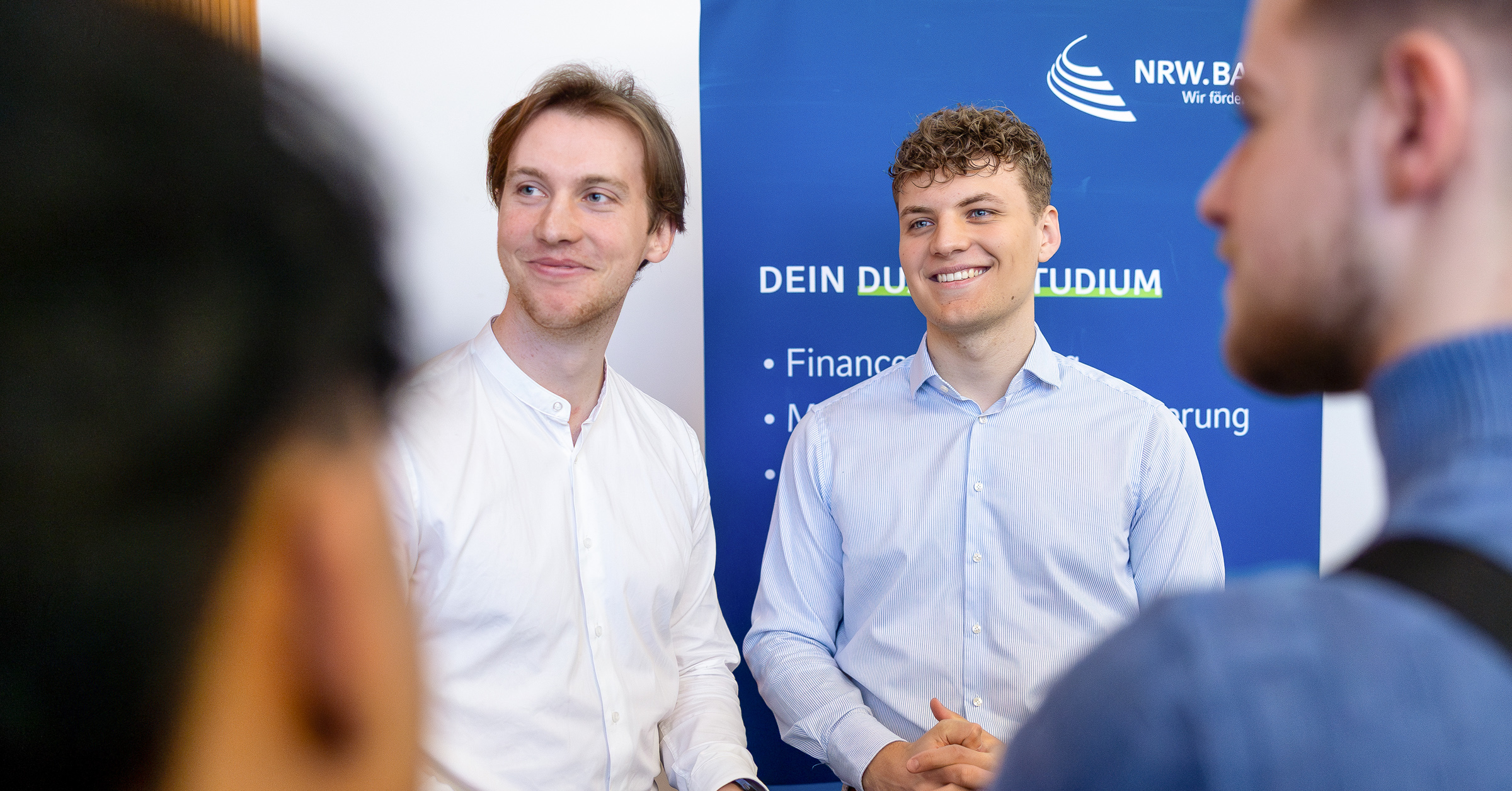 Zwei junge Männer in weißen Hemden stehen an einem Infostand der NRW.BANK bei einer Veranstaltung der Ausbildungsoffensive. Sie lächeln und führen ein Gespräch mit zwei Jugendlichen. Im Hintergrund ist ein blaues Roll-up mit Informationen zum dualen Studium sichtbar.