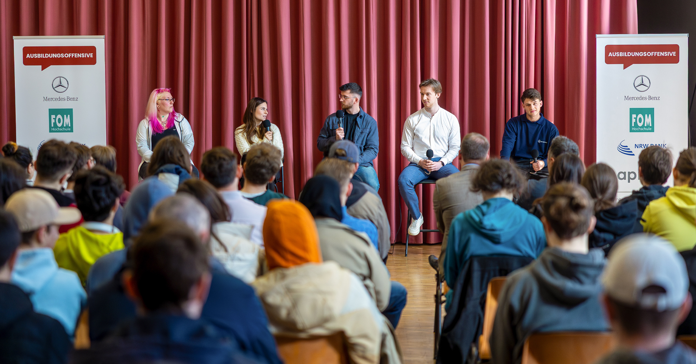 Fünf junge Erwachsene sitzen bei einer Talkrunde der Ausbildungsoffensive vor einem roten Vorhang auf einer Bühne und sprechen mit Mikrofonen über ihre Ausbildungserfahrungen. Das Publikum besteht aus zahlreichen Jugendlichen. Zwei Roll-ups mit Logos von Partnern wie Mercedes-Benz, FOM Hochschule, NRW.BANK und apm flankieren die Bühne.