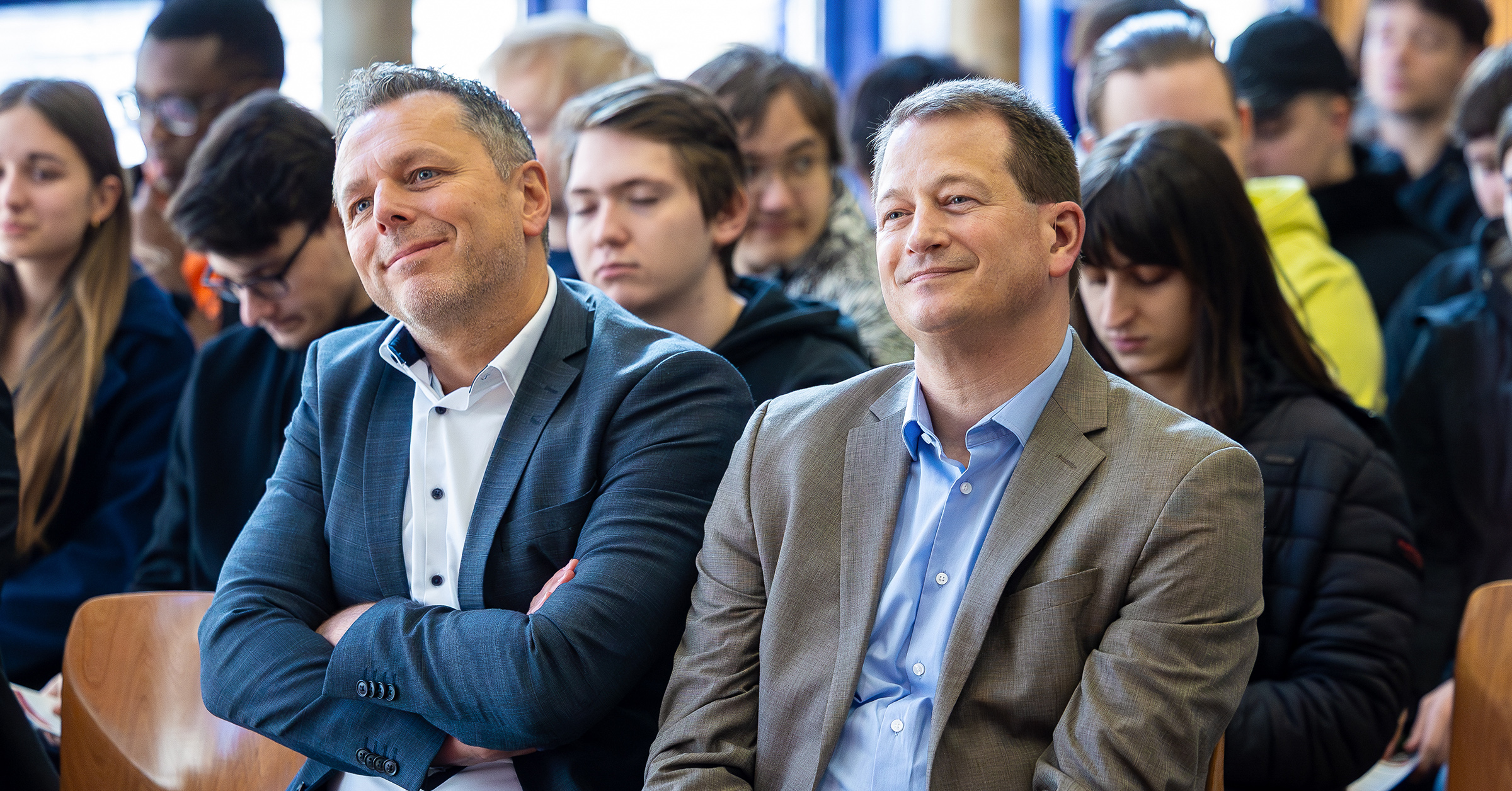 Zwei Männer in Anzügen sitzen in der ersten Reihe einer Veranstaltung der Ausbildungsoffensive und blicken mit freundlichen Gesichtern zur Bühne. Hinter ihnen folgen weitere Teilnehmende – überwiegend SchülerInnen – aufmerksam dem Geschehen.