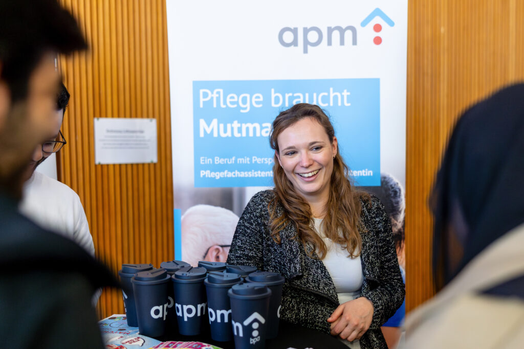 Eine ausbildungsverantwortliche Mitarbeiterin von APM am Infostand des Untenehmens bei einem Schulbesuch der Ausbildungsoffensive in Düsseldorf. Die Frau lächelt im Gespräch mit mehreren Jugendlichen. Im Hintergrund ist ein Roll-Up des Ausbildungspartners APM mit dem Slogan ‚Pflege braucht Mutmacher‘ zu sehen. Auf dem Tisch stehen mehrere blaue Mehrwegbecher mit dem APM-Logo. Die Szene spielt sich in einer Schule ab und zeigt lebendige Berufsorientierung auf Augenhöhe.